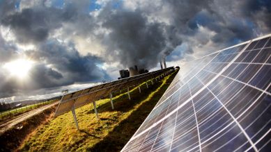 Die Sonne scheint auf eine Solaranlage, im Hintergrund ein Kraftwerk in den Wolken © picture alliance/ dpa/ Oliver Berg