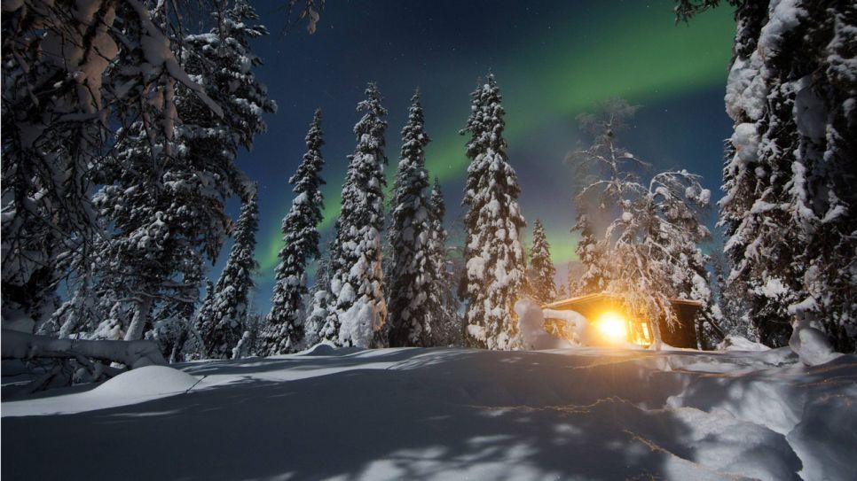 Eine beleuchtete Hütte im Winterwald, darüber Polarlichter (Quelle: imago/Frank Sorge)